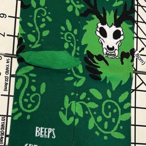 Puede incluir: Calcetines verdes con un gráfico de calavera y cuernos blancos, rodeados de un diseño de hojas verdes. El texto "BEEPS CREATURES" está en blanco. Los calcetines tienen punteras y puños verdes.