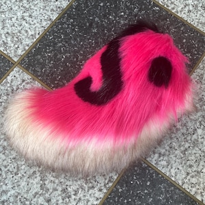 May include: A bright pink faux fur tail with black markings. The tail has a gradient from white to pink, with a black swirl design. The fluffy tail is designed for cosplay or costume use.