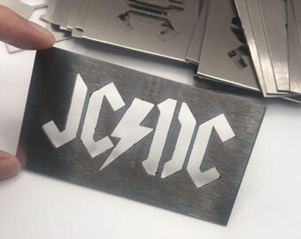 May include: A collection of metal stencils featuring the band name "AC/DC" in a bold, stylized font with a lightning bolt. The stencils are a dark gray color and appear to be made of brushed metal. The stencils are rectangular.