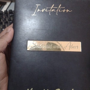 May include: Black invitation card with gold lettering. The card says "Invitation" at the top, "Shukraib wed Abeer" in the middle, and "Mr & Mrs Raheel" at the bottom.