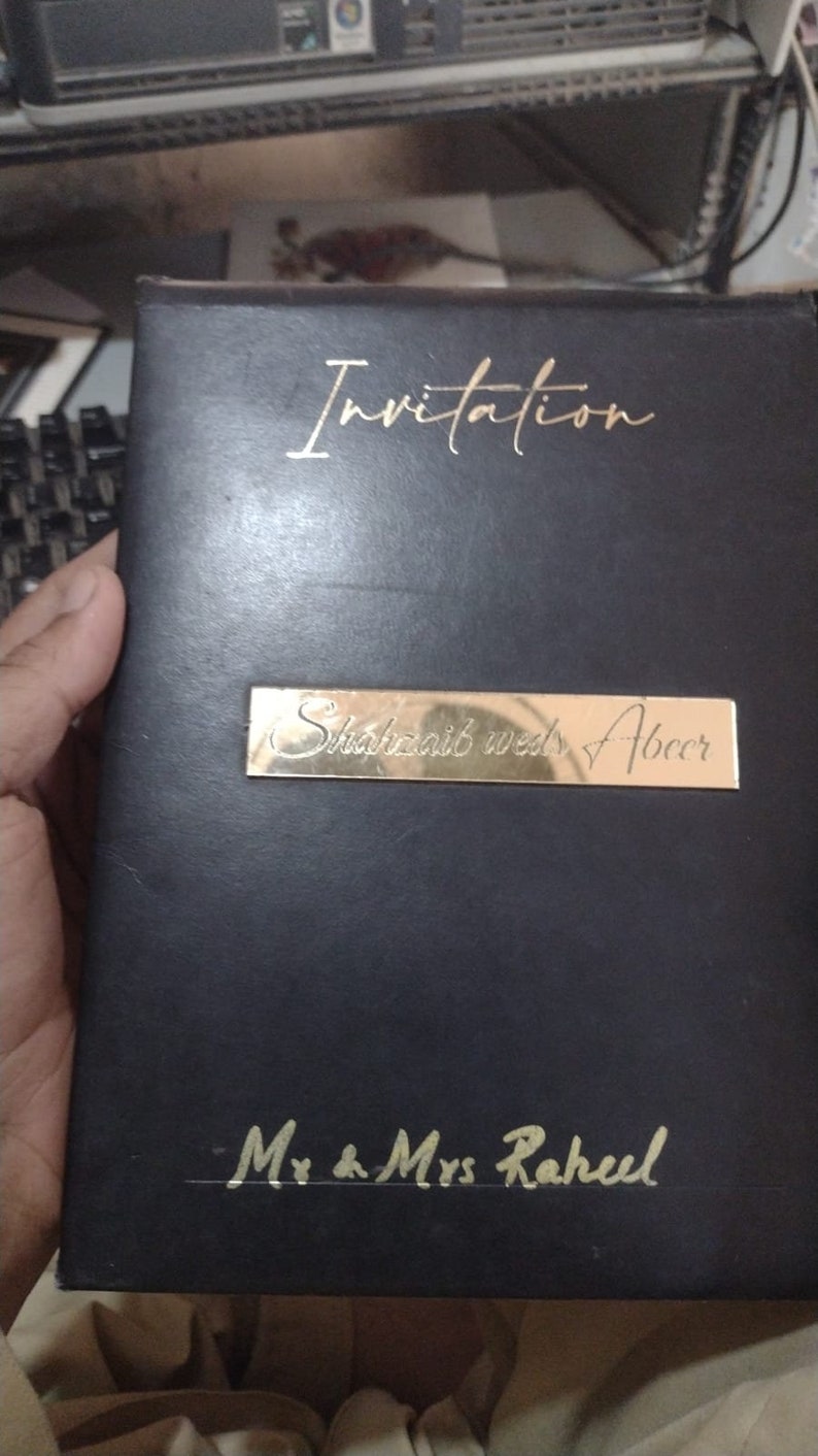 May include: Black invitation card with gold lettering. The card says "Invitation" at the top, "Shukraib wed Abeer" in the middle, and "Mr & Mrs Raheel" at the bottom.