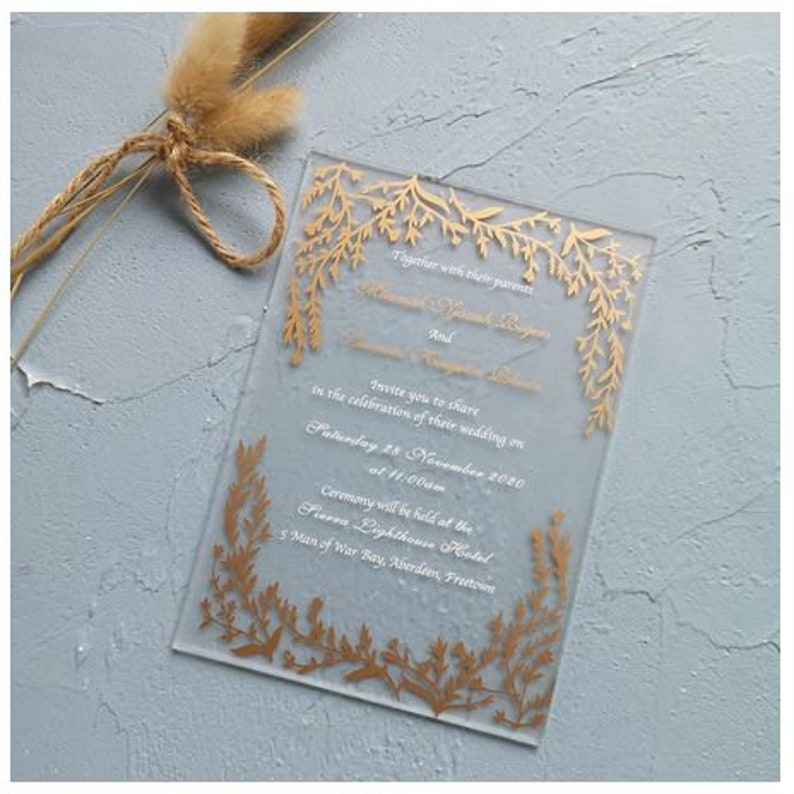 May include: A clear acrylic wedding invitation with gold foil floral accents. The invitation text reads "Together with their parents, [Name] and [Name], invite you to share in the celebration of their wedding on Saturday 28 November 2020 at 11:00am. Ceremony will be held at the Fierro Lighthouse Hotel, 5 Man of War Bay, Aberdeen, Fretteson."