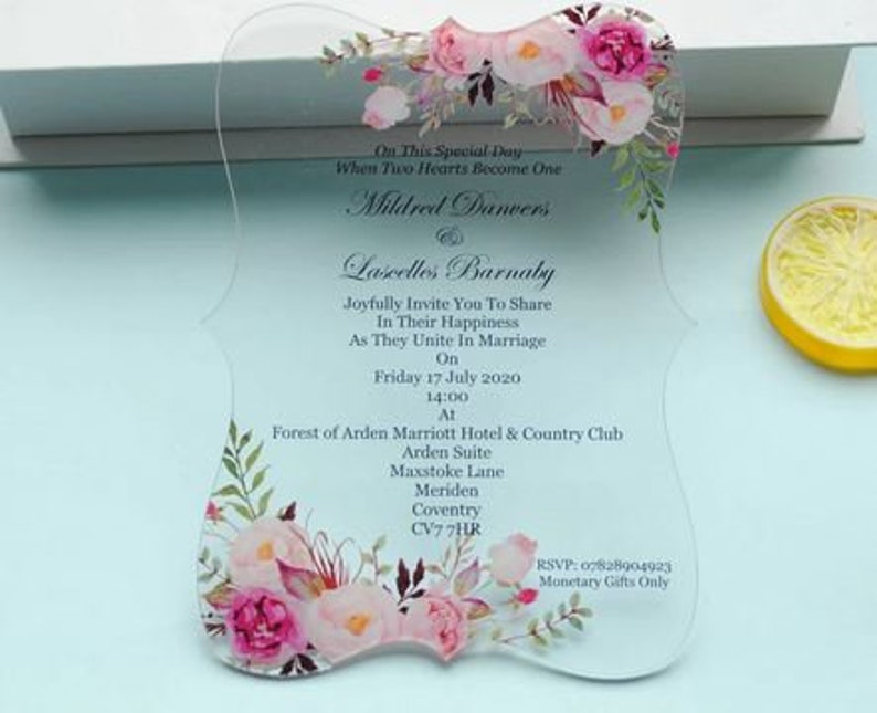May include: Clear acrylic wedding invitation with pink and white floral design. The invitation reads "On This Special Day When Two Hearts Become One Mildred Danvers & Lascelles Barnaby Joyfully Invite You To Share In Their Happiness As They Unite In Marriage On Friday 17 July 2020 14:00 At Forest of Arden Marriott Hotel & Country Club Arden Suite Maxstoke Lane Meriden Coventry CV7 7HR RSVP: 07828904923 Monetary Gifts Only"