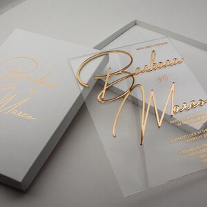 May include: Clear acrylic wedding invitation with gold foil lettering. The invitation reads "With great pleasure Barbara & Marco invites you to join them at the celebration of their marriage at ten o'clock in the morning, Saturday, June 10th, PortMiami-Cruise Terminal 1015 North America Way, Miami, Caribbean".