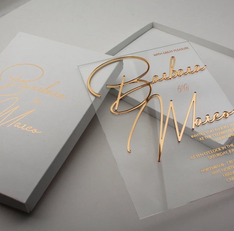 May include: Clear acrylic wedding invitation with gold foil lettering. The invitation reads "With great pleasure Barbara & Marco invites you to join them at the celebration of their marriage at ten o'clock in the morning, Saturday, June 10th, PortMiami-Cruise Terminal 1015 North America Way, Miami, Caribbean".