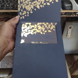 May include: A navy blue wedding invitation with a gold foil design of leaves. The invitation has a clear pocket on the front to hold a smaller card with the wedding details. The text on the smaller card reads "The honour of your presence is requested at the wedding of Mr. [Name] and Ms. [Name]"