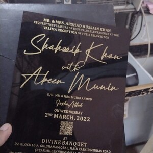 May include: Black and gold wedding invitation card with the names Shakhsaik Khan and Abeer Munir. The invitation is for a Valima reception on Wednesday, March 2nd, 2022 at Divine Banquet in Karachi, Pakistan. The invitation includes contact information and details about the gathering and dinner times.