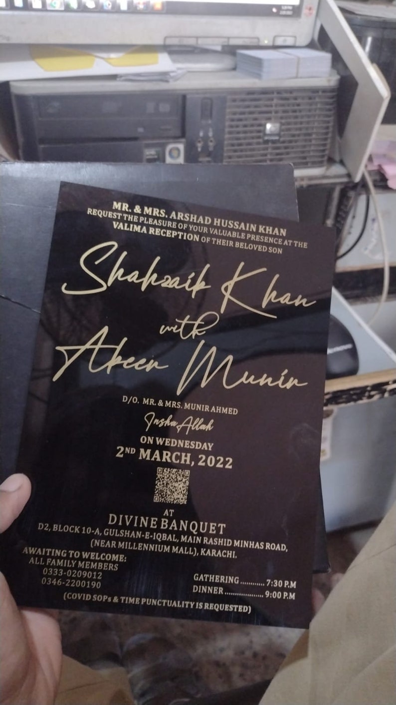 May include: Black and gold wedding invitation card with the names Shakhsaik Khan and Abeer Munir. The invitation is for a Valima reception on Wednesday, March 2nd, 2022 at Divine Banquet in Karachi, Pakistan. The invitation includes contact information and details about the gathering and dinner times.