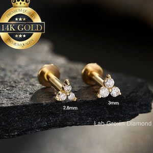 May include: Two gold-colored labret earrings with a cluster of three clear, round diamonds. One earring is 2.8mm, the other is 3mm. The earrings are displayed on a dark, textured surface. The image includes a gold seal that says "Premium Quality 14K Gold".