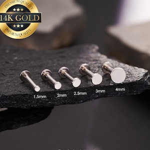 以下が含まれることがあります： 1.5mmから4mmまでのサイズの、銀色のフラットバックラブレットスタッドが5つ、暗く質感のある表面に表示されています。左上には「14K GOLD」の文字が入った金のエンブレムがあります。「@ouferbodyjewelry」のInstagramハンドルが下にあります。