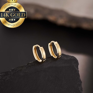 May include: A pair of gold-coloured hoop earrings. The earrings have a small, flat, rectangular design. The image includes a gold seal that reads "Premium Quality" and "14K Gold". The earrings are displayed on a dark, textured surface.