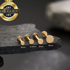 Peut inclure: Quatre boucles d'oreilles dorées à dos plat avec des faces circulaires texturées, allant de 2 mm à 4 mm. Les boucles d'oreilles sont présentées sur une surface sombre et texturée. Un sceau doré avec le texte "14K GOLD" est en haut à gauche.