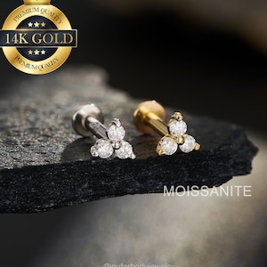 Könnte beinhalten: Zwei Ohrstecker, einer silberfarben und einer goldfarben, jeweils mit einem Cluster aus drei klaren, runden Edelsteinen. Der goldfarbene Stecker ist mit "14K GOLD" und "MOISSANITE" beschriftet. Der Schmuck wird auf einer dunklen, strukturierten Oberfläche präsentiert.