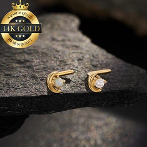 May include: A pair of gold-colored stud earrings, each featuring a crescent moon design. One earring has a white gemstone, and the other has a clear, sparkling gemstone. The earrings are displayed on a dark, textured surface. The image includes a gold seal that says "14K GOLD".