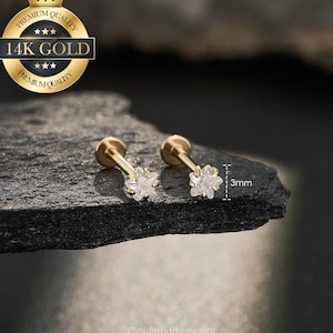 May include: Two gold-colored, flower-shaped earrings with clear gemstones. The earrings are 3mm in size and are displayed on a dark, textured surface. The image includes a gold seal that reads "Premium Quality" and "14K Gold".