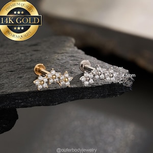 May include: Two flower-shaped ear studs. One is gold-toned, the other silver-toned, each with three flower clusters. The flowers have clear, sparkling accents. The gold stud is marked "14K GOLD". The studs are on a dark, textured surface.
