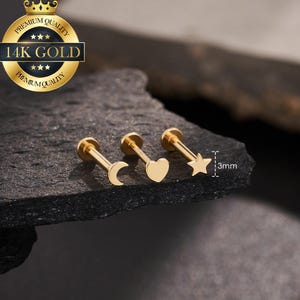 May include: Three gold-colored lip or cartilage studs with a moon, heart, and star design. The studs are displayed on a dark, textured surface. The image includes a gold seal that reads "14K GOLD". The text "3mm" is also visible.