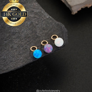 Puede incluir: Tres pequeños colgantes redondos de ópalo en azul, morado y blanco, cada uno con un lazo dorado. Los colgantes se exhiben sobre una superficie oscura y texturizada. Un emblema dorado dice "Premium Quality" y "14K Gold". Se ve el identificador de Instagram @ouferbodyjewelry.