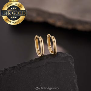 May include: A pair of gold-colored oval hoop earrings. The earrings are displayed on a dark, textured surface. A gold seal in the upper left corner reads "Premium Quality" and "14K Gold". The Instagram handle "@ouferbodyjewelry" is in the lower right corner.