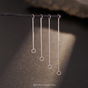 Pode incluir: Quatro correntes de metal prateado de comprimentos variados penduradas num fio horizontal preto fino. Cada corrente apresenta um pequeno anel de junção no topo e um anel maior na parte inferior. O texto @ouferbodyjewelry é visível.