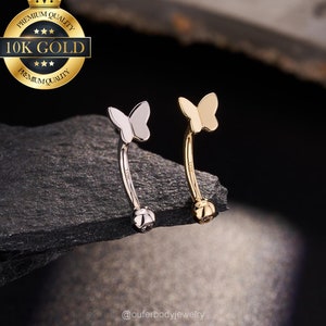 May include: Two curved eyebrow rings with butterfly designs. One is silver-colored, the other gold-colored. The jewelry is displayed on a dark, textured surface. The image includes a gold seal that says "Premium Quality 10K Gold".
