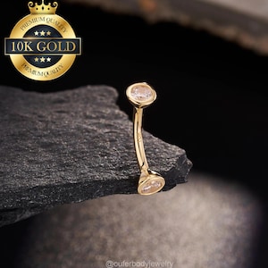 May include: Gold body jewelry with two clear gemstones. The jewelry is curved and has a 10K Gold label.