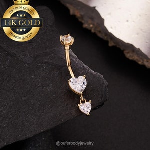 May include: A gold-colored belly button ring with a curved design. The ring features a clear gemstone at the top and two heart-shaped gemstones dangling below. The words "14K GOLD" are printed on a gold-colored emblem.