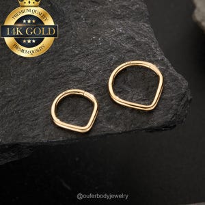 Puede incluir: Dos pendientes dorados de forma geométrica. Los pendientes se muestran sobre una superficie oscura y texturizada. Un sello dorado con el texto "14K GOLD" está en la esquina superior izquierda. Los pendientes tienen un diseño articulado.