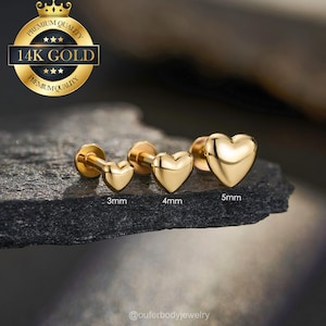 May include: Three gold heart-shaped earrings in 3mm, 4mm, and 5mm sizes. The earrings are displayed on a dark, textured surface. A gold emblem with the text "14K GOLD" is in the upper left corner.