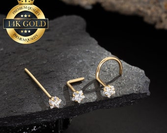 Puede incluir: Tres anillos de nariz de oro con un pequeño diseño de flor de cristal transparente. Los anillos están sobre un fondo gris oscuro. El texto "14K GOLD" está en un círculo dorado con una corona en la parte superior.