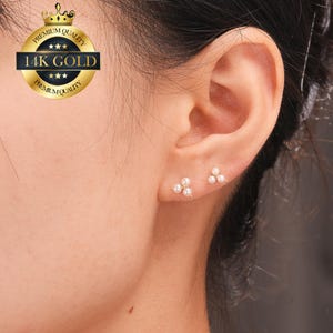 May include: Close-up of two pairs of gold stud earrings, each featuring three small, white pearls arranged in a triangular cluster. The earrings are set against the backdrop of a person's ear, showcasing the jewelry's delicate design and elegant simplicity.