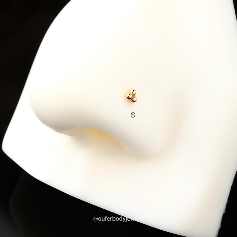 Puede incluir: Anillo de nariz de oro con tres peque&ntilde;as cuentas redondas. El anillo est&aacute; en un expositor blanco. El texto "S" est&aacute; debajo del anillo.