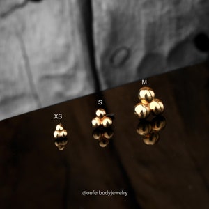 Puede incluir: Tres tama&ntilde;os de joyer&iacute;a de cuerpo de racimo de oro, etiquetados como XS, S y M. La joyer&iacute;a est&aacute; hecha de tres peque&ntilde;as bolas agrupadas.