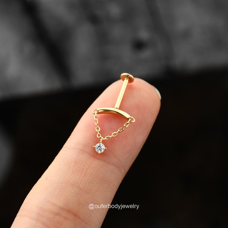May include: Gold body jewelry with a small diamond charm hanging from a chain. The chain is attached to a curved bar that goes through the piercing.