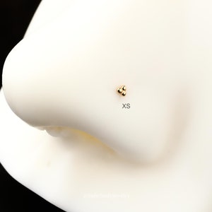 Puede incluir: Anillo de nariz de oro con tres peque&ntilde;as bolas. El anillo est&aacute; en una nariz de maniqu&iacute; blanca. El texto "XS" est&aacute; debajo del anillo.