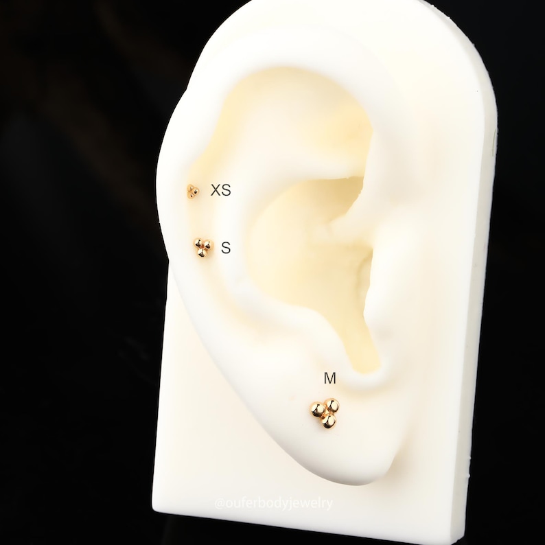 Puede incluir: Primer plano de una oreja modelo con tres tama&ntilde;os diferentes de pendientes de bola de oro. Los pendientes est&aacute;n etiquetados como "XS", "S" y "M".