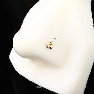 Puede incluir: Anillo de nariz de oro con tres peque&ntilde;as cuentas redondas en forma de tri&aacute;ngulo. El anillo est&aacute; en una nariz de maniqu&iacute; blanca.