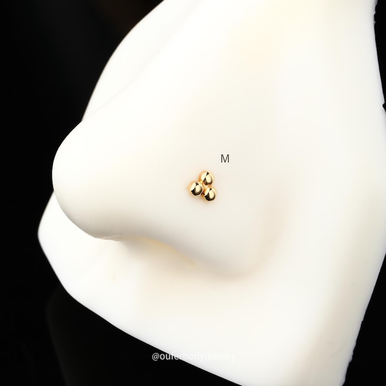 Puede incluir: Anillo de nariz de oro con tres peque&ntilde;as cuentas redondas en forma de tri&aacute;ngulo. El anillo est&aacute; en una nariz de maniqu&iacute; blanca.