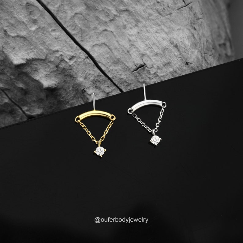 May include: Two minimalist earrings with a small diamond-shaped stone hanging from a delicate chain. The earrings are available in gold and silver.