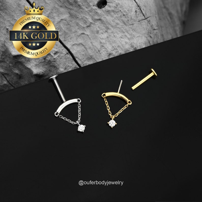 May include: A pair of 14k gold body jewelry pieces with a curved bar and a dangling chain with a small clear stone. The jewelry is on a black background with a gold and white label that says "Premium Quality 14k Gold Premium Quality".