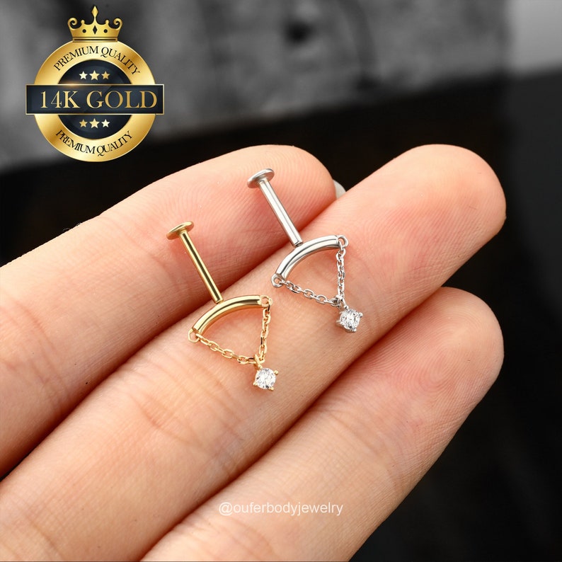May include: Two 14k gold body jewelry pieces with a curved bar and a dangling chain with a small diamond at the end. The jewelry is shown on a hand.