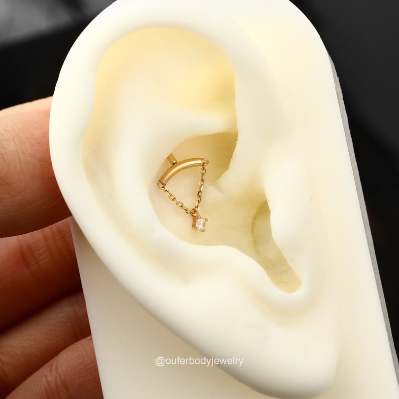 May include: Gold ear piercing with a small diamond dangle and a curved bar with a chain connecting the two pieces. The piercing is in a model's ear.