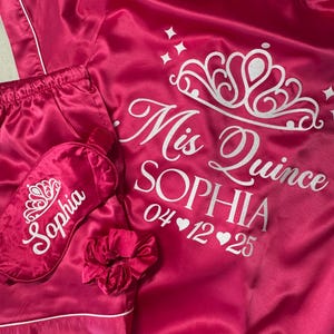 May include: Pink satin robe and shorts set with a white crown and the text "Mis Quince Sophia 04♥12♥25". The set also includes a pink satin sleep mask with a white crown and the text "Sophia".