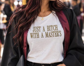 May include: A white t-shirt featuring the gold text JUST A BITCH WITH A MASTER'S, worn under a black graduation gown with maroon trim, paired with black jeans, a black graduation cap with a gold tassel, and a black diploma holder.