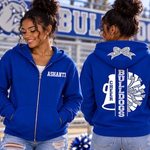 May include: A royal blue zip-up hoodie featuring the name ASHANTI in white on the front left chest. The back displays a silver glitter cheer bow, a white megaphone, the word CHEER, and the text BULLDOGS with a silver glitter pom-pom graphic.