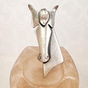 Op de afbeelding: Een zilverkleurige metalen sculptuur van een paardenkop, met een gestileerd ontwerp, puntige oren en een gedetailleerde manen. De sculptuur is gemonteerd op een mat, perzikroze voet, wat een opvallend contrast creëert. Een decoratief kunstwerk.