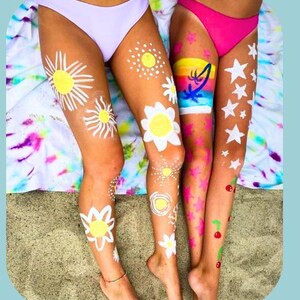 Colorful body paint designs on their legs, featuring suns, stars, rainbows, flowers, and cherries, drawn with face and body paint crayons. Perfect for summer, beach days, or party fun