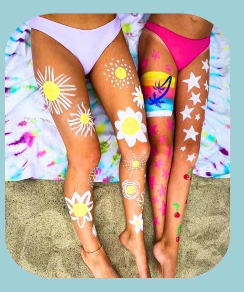 Colorful body paint designs on their legs, featuring suns, stars, rainbows, flowers, and cherries, drawn with face and body paint crayons. Perfect for summer, beach days, or party fun