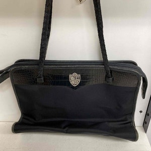 K&ouml;nnte beinhalten: Eine schwarze Ledertasche mit geflochtenem Trageriemen und einem silbernen Herz-Anh&auml;nger. Die Tasche hat ein Kroko-Pr&auml;gemuster und ein silbernes, schildf&ouml;rmiges Logo.