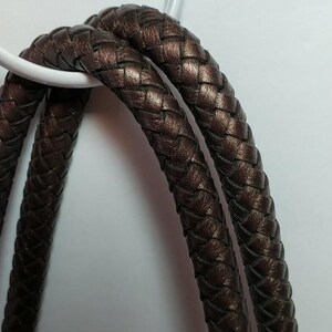 May include: Close-up of a brown braided leather strap, looped over a white circular object. The leather has a metallic sheen. The background is a plain white surface. The strap appears to be part of a bag or accessory.